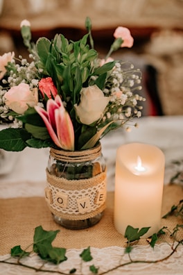 A decorative arrangement featuring a glass jar adorned with a lace and burlap wrap, labeled with wooden Scrabble tiles spelling 'LOVE'. Inside the jar is a bouquet of mixed flowers, including roses and lilies, with greenery accents. Next to the jar, a lit pillar candle casts a warm glow, all set on a lace tablecloth with vine elements.