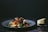 A plate of pasta garnished with cherry tomatoes, olives, and herbs sits on a dark surface. Next to the plate, a wedge of cheese is placed on a small cutting board.
