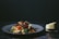 A plate of pasta garnished with cherry tomatoes, olives, and herbs sits on a dark surface. Next to the plate, a wedge of cheese is placed on a small cutting board.