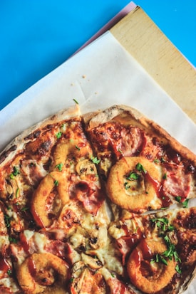 A close-up view of a pizza featuring a variety of toppings, including fried onion rings, mushrooms, ham, melted cheese, and herbs. The pizza is partially sliced and set on parchment paper atop a wooden board with a blue background.