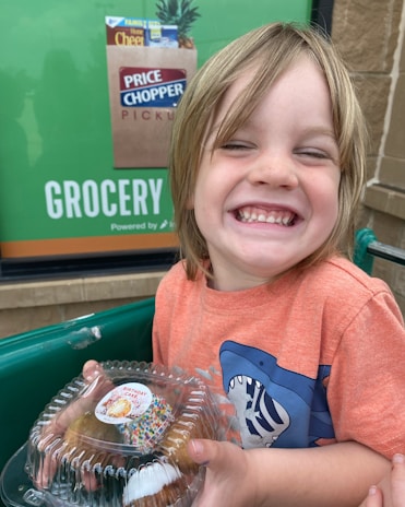 A happy child with long hair and a big smile is holding a clear plastic container with a birthday cake inside. The child is wearing an orange T-shirt with a shark design. In the background, there is a poster advertising Price Chopper grocery pickup services.