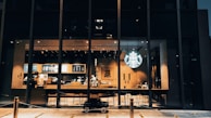 A modern coffee shop with large glass windows displaying the interior seating area and the counter. The iconic logo is illuminated, and the warm lighting creates a cozy atmosphere. Two employees are visible behind the counter, with stools lined up in front. A small toy car is parked on the pavement outside.