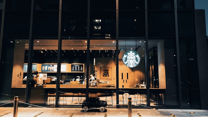 A modern coffee shop with large glass windows displaying the interior seating area and the counter. The iconic logo is illuminated, and the warm lighting creates a cozy atmosphere. Two employees are visible behind the counter, with stools lined up in front. A small toy car is parked on the pavement outside.
