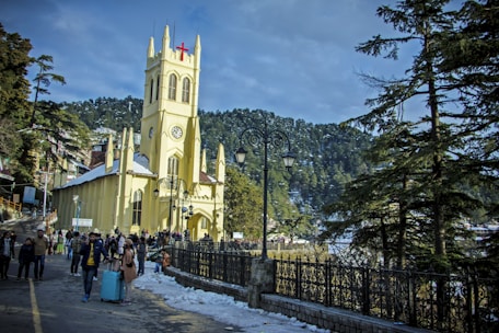 A Gothic-style church with a tall steeple and a prominent red cross is surrounded by snow on the ground. People are walking along a pathway next to the church, and a lamppost stands nearby. Tall pine trees and hills provide a scenic backdrop, under a partly cloudy sky.