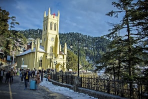 A Gothic-style church with a tall steeple and a prominent red cross is surrounded by snow on the ground. People are walking along a pathway next to the church, and a lamppost stands nearby. Tall pine trees and hills provide a scenic backdrop, under a partly cloudy sky.