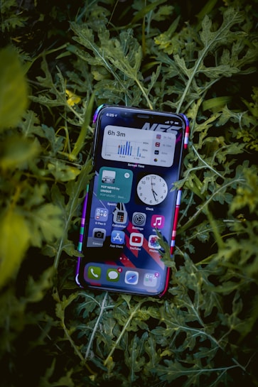 A smartphone displaying a variety of apps is resting on a bed of green leaves. The screen shows multiple applications, including music, messaging, and time tracking, all organized neatly.
