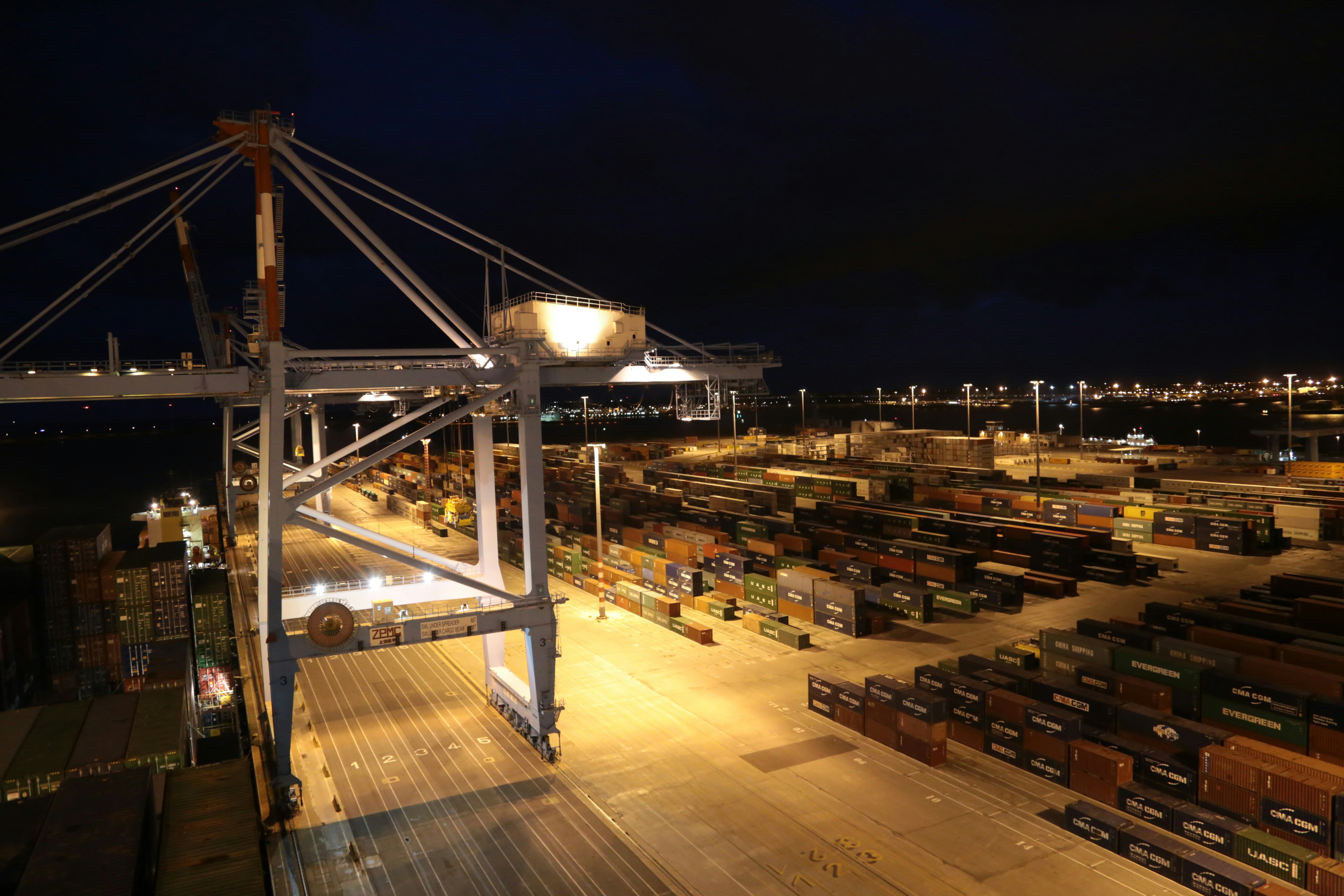 Cargo containers stacked at night in a busy port, dark industrial atmosphere with orange crane lights