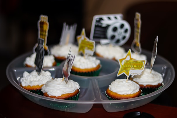 Cupcakes with white frosting are arranged on a tray, each topped with movie-themed decorations such as film reels, stars, and clapperboards.