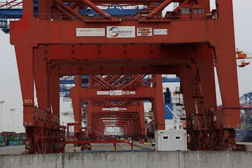 Rows of large red cranes dominate the scene within an industrial shipping port. The cranes tower over a road with a small office building visible on the right side. Containers and additional cranes can be seen in the background.