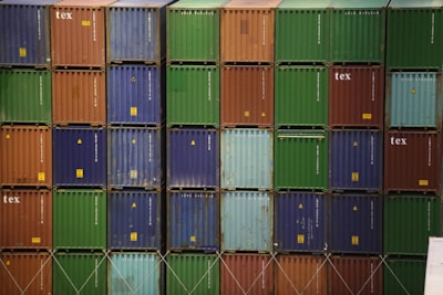 A stacked arrangement of colorful shipping containers with visible text on some of them. The containers are organized in a grid pattern, showcasing various colors including green, blue, red, and orange.