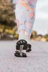 Close-up view of a person's legs in patterned leggings and running shoes walking on a paved path. The background appears blurred with greenery, indicating an outdoor setting.