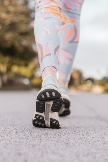 Close-up view of a person's legs in patterned leggings and running shoes walking on a paved path. The background appears blurred with greenery, indicating an outdoor setting.