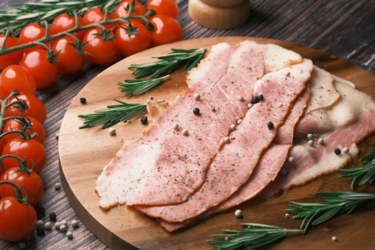 Thin slices of cured meat are arranged on a wooden board, garnished with sprigs of rosemary and scattered peppercorns. A bunch of ripe cherry tomatoes on the vine is placed nearby, adding a burst of red to the composition. The wooden surface beneath adds a rustic touch.