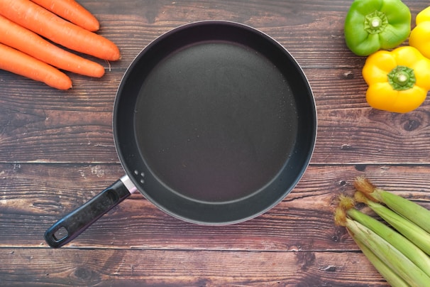 Close-up of colorful non-stick frying pans stacked with fresh vegetables beside them.
