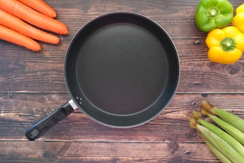 A black frying pan is placed on a wooden surface surrounded by fresh vegetables, including three orange carrots on the left, a green bell pepper and yellow bell peppers on the right, and corn cobs on the lower right corner.