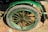 A close-up view of a green bicycle or cart wheel with a tire mounted on a metal rim. The wheel features multiple spokes and has a surrounding fender. The image is taken from a top angle, showing part of the brick pavement underneath.