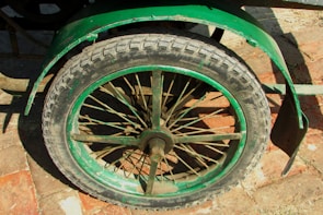 A close-up view of a green bicycle or cart wheel with a tire mounted on a metal rim. The wheel features multiple spokes and has a surrounding fender. The image is taken from a top angle, showing part of the brick pavement underneath.