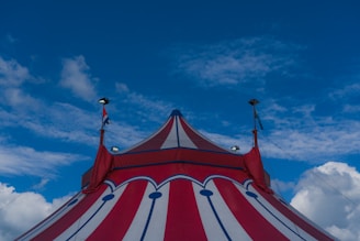 A colorful circus tent with red and white stripes extends under a bright blue sky. Small flags are visible near the top of the tent, and the structure is held up by supporting ropes.