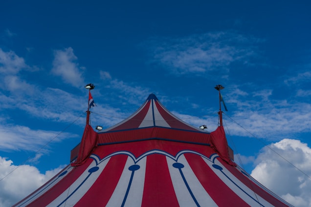 A colorful circus tent with red and white stripes extends under a bright blue sky. Small flags are visible near the top of the tent, and the structure is held up by supporting ropes.