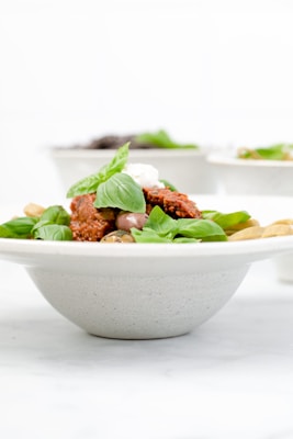 A bowl filled with fresh basil leaves, dollops of creamy white sauce, and a rich red tomato-based sauce, possibly pasta or a similar dish. The focus is on the vibrant colors and textures of the ingredients.