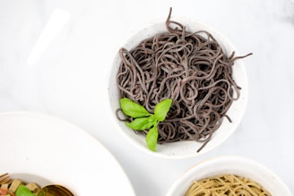 A bowl of dark-colored noodles garnished with fresh green basil leaves. The bowl is placed on a white surface, and partially visible are other bowls containing different types of noodles and food items.