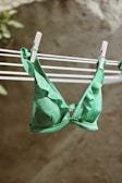 A green bikini top with ruffled edges is hanging on a clothesline secured by white clothespins. The background is blurred, with a hint of greenery on the left.