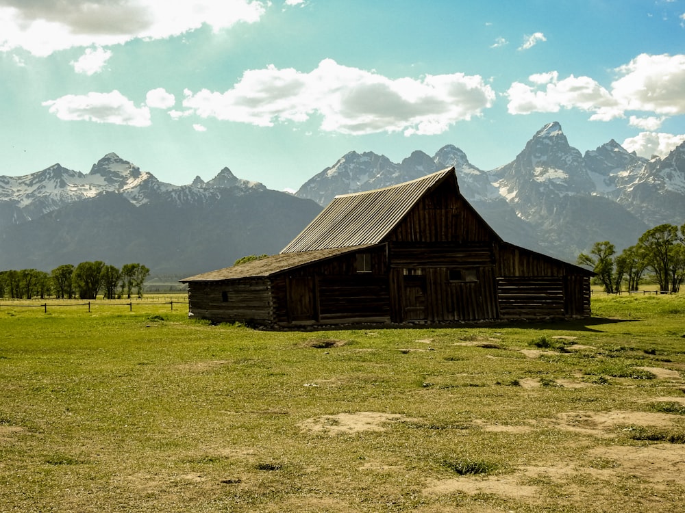 grange en bois marron sur un champ d’herbe verte près d’une montagne enneigée sous des nuages blancs et bleus