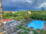 A serene outdoor setting featuring a hotel pool area, surrounded by lush greenery and palm trees. There are sun loungers with thatched umbrellas arranged neatly along the poolside, and a seating area with white tables and chairs under pergolas. The sky is overcast, suggesting an impending storm.