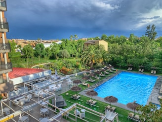 A serene outdoor setting featuring a hotel pool area, surrounded by lush greenery and palm trees. There are sun loungers with thatched umbrellas arranged neatly along the poolside, and a seating area with white tables and chairs under pergolas. The sky is overcast, suggesting an impending storm.