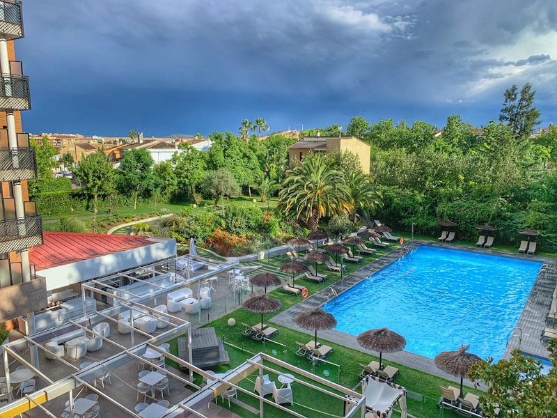 A serene outdoor setting featuring a hotel pool area, surrounded by lush greenery and palm trees. There are sun loungers with thatched umbrellas arranged neatly along the poolside, and a seating area with white tables and chairs under pergolas. The sky is overcast, suggesting an impending storm.