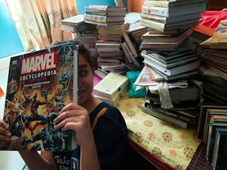 A person holds up a large Marvel Encyclopedia book in front of their face, with stacks of various books piled high on a table in the background. The room is well-lit by natural light coming through a window with drapes.