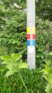 A concrete pole with colorful painted squares and rectangles in yellow, red, and blue against a lush green background of leaves and plants.