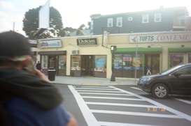 A street view featuring a crosswalk leading to a row of commercial buildings. The businesses visible include a restaurant and a convenience store. A person with a blurred face stands in the foreground, while a car is seen to the right. The sky is slightly overcast, and a billboard structure is seen above the buildings.