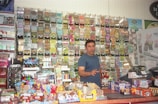 A person stands behind the counter of a convenience store, surrounded by a wall of scratch-off lottery tickets, various candy items, and snacks. The counter is filled with brightly colored boxes and display items.