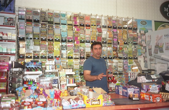 A person stands behind the counter of a convenience store, surrounded by a wall of scratch-off lottery tickets, various candy items, and snacks. The counter is filled with brightly colored boxes and display items.