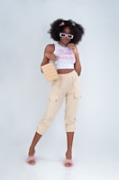 A stylish woman stands confidently, wearing sunglasses, a white crop top with the words 'Bad Habits', and beige pants. She carries a square-shaped handbag and wears fluffy pink slippers. Her hair is voluminous and decorated with colorful clips.