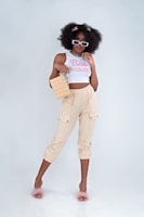 A stylish woman stands confidently, wearing sunglasses, a white crop top with the words 'Bad Habits', and beige pants. She carries a square-shaped handbag and wears fluffy pink slippers. Her hair is voluminous and decorated with colorful clips.
