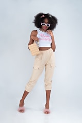 A stylish woman stands confidently, wearing sunglasses, a white crop top with the words 'Bad Habits', and beige pants. She carries a square-shaped handbag and wears fluffy pink slippers. Her hair is voluminous and decorated with colorful clips.