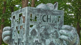 A bronze statue of a cartoonish cat is holding a book or sign with cut-out letters spelling 'LE CHAT'. The statue has a playful and artistic design, with large round ears and oversized hands. It is set against a backdrop of lush green trees, suggesting a park or garden setting.