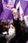 A group of people participate in a protest with raised fists, wearing face masks that carry messages. Purple flags with various symbols and text are prominently visible, symbolizing solidarity and activism.