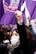 A group of people participate in a protest with raised fists, wearing face masks that carry messages. Purple flags with various symbols and text are prominently visible, symbolizing solidarity and activism.