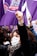 A group of people participate in a protest with raised fists, wearing face masks that carry messages. Purple flags with various symbols and text are prominently visible, symbolizing solidarity and activism.