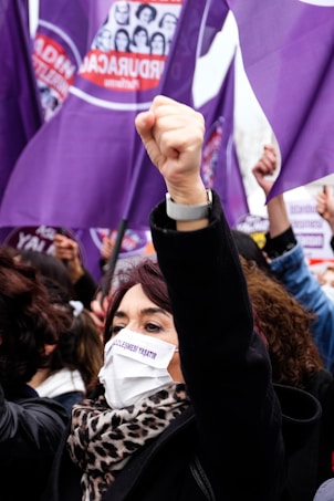 A group of people participate in a protest with raised fists, wearing face masks that carry messages. Purple flags with various symbols and text are prominently visible, symbolizing solidarity and activism.
