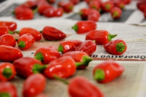 Red peppers are scattered across newspaper pages. The focus is on the peppers, with their shiny, smooth texture and vibrant green stems standing out against the monochrome background of the newspaper.