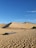 A vast desert landscape with large sand dunes under a clear blue sky. There are people walking and riding camels across the desert. In the foreground, a rectangular structure and a circular object are present on the sandy ground, along with a patch of green vegetation to the right.