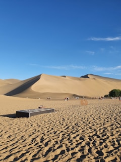 A vast desert landscape with large sand dunes under a clear blue sky. There are people walking and riding camels across the desert. In the foreground, a rectangular structure and a circular object are present on the sandy ground, along with a patch of green vegetation to the right.