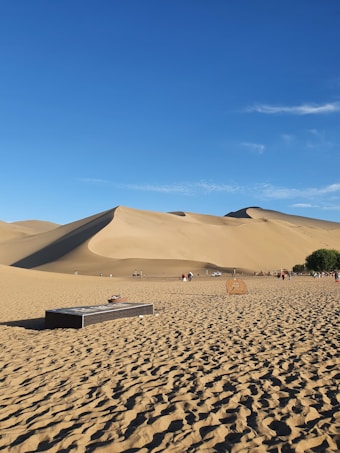 A vast desert landscape with large sand dunes under a clear blue sky. There are people walking and riding camels across the desert. In the foreground, a rectangular structure and a circular object are present on the sandy ground, along with a patch of green vegetation to the right.