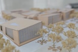 A detailed architectural model features several modern buildings arranged in a complex. The structures have geometric designs with large windows and beige exteriors. Scale model trees with light foliage are interspersed between the buildings, all placed on a grid-patterned surface that simulates an urban planning layout.