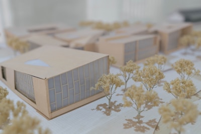 A detailed architectural model features several modern buildings arranged in a complex. The structures have geometric designs with large windows and beige exteriors. Scale model trees with light foliage are interspersed between the buildings, all placed on a grid-patterned surface that simulates an urban planning layout.