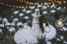 A softly lit outdoor wedding reception setup with elegant tables, delicate floral arrangements, and string lights under a twilight sky.